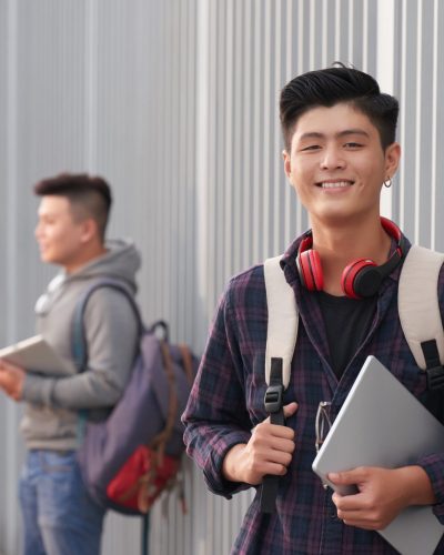 Waist-up portrait of handsome Asian student posing for photography with wide smile while standing at facade of university building